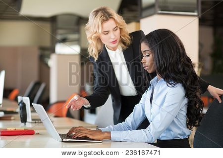 Africam American Woman Sitting At Desk. Er White Colleague Standing Nearby.