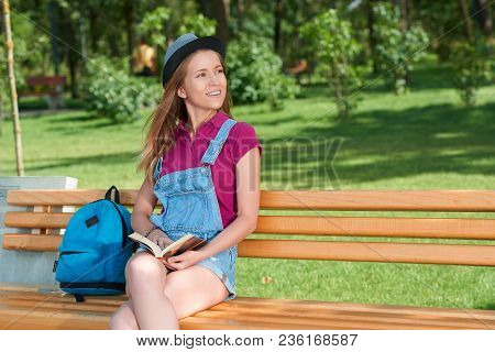 Young Pretty Girl Sitting On The Benchin The City Park Keeping A Book. Wearing Blue Jeans Shorts, Br