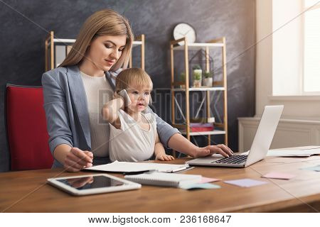 Happy Beautiful Business Mom Working On Laptop In Office While Her Cute Baby Holding Smartphone. Bus