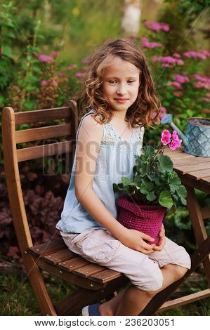 Dreamy Happy Child Girl Relaxing In Summer Evening Garden With Heranium Flower In Pot. Teaching Kids