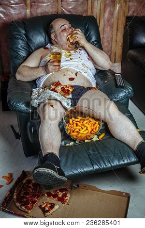 Photo Of A Fat Couch Potato Eating A Huge Hamburger And Watching Television.  Harsh Lighting From Th