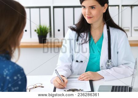 Beautiful Female Doctor And Patient Having A Talk While Sitting At The Desk. Physician Writes Prescr