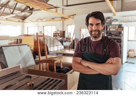 Portrait Of A Smiling Young Craftsman Standing With His Arms Crossed By A Bench Saw In His Workshop 