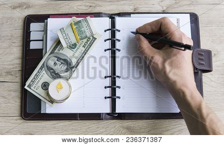 Ethereum Crypto Currency On Top Dollar Biils On Notepad With Mans Hand With Pen. Profit From Mining 