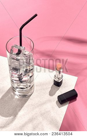 High Angle View Of Lipstick And Drinking Glass With Ice On Table. Pink And White Background With Sha