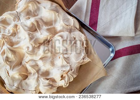 Cooking the meringue base for a Pavlova dessert. Fresh meringue, with crisp golden top, on baking parchment and a stainless steel baking tray. Overhead view on wooden table with cloth.