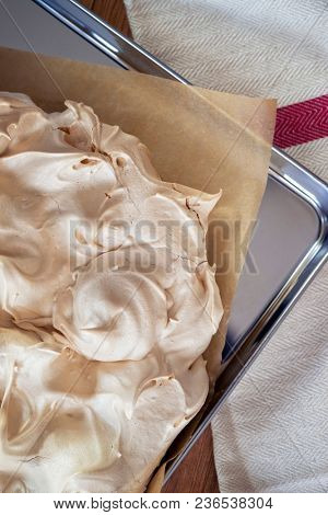 Cooking the meringue base for a Pavlova dessert. Fresh meringue, with crisp golden top, on baking parchment and a stainless steel baking tray. Overhead view on wooden table with cloth.