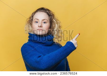 Check This Out. Serious Blonde Young Woman With Curly Blonde Hair Dressed In Bright Blue Sweater, In