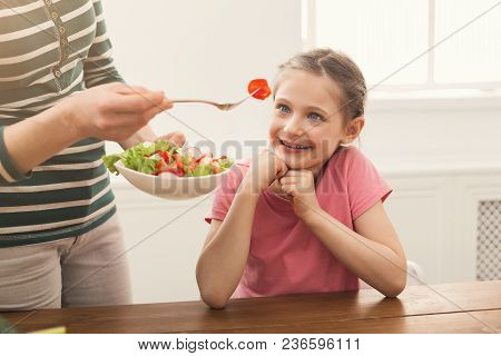 Woman Offering Her Happy Daughter Salad In The Kitchen Over Window. Healthy Food For Children Concep