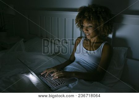 Young casual woman lounging on bed in dark room in nigh time using laptop and typing while communicating in virtual world.