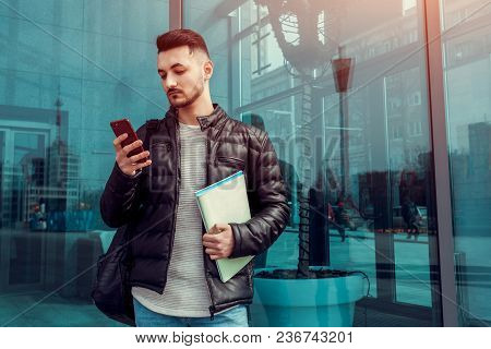 Confident Arabian Student Using Smartphone Outside. Serious Guy Looks At Phone In Front Of Modern Bu