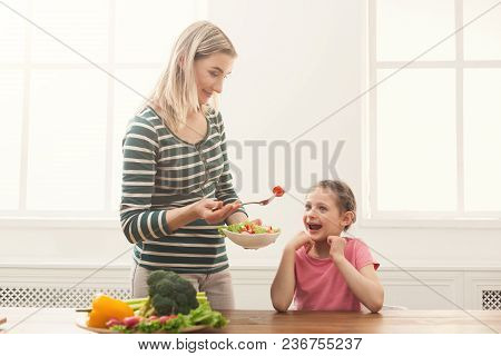 Beautiful Woman Offering Her Happy Daughter Salad In The Kitchen Over Window. Healthy Food For Child