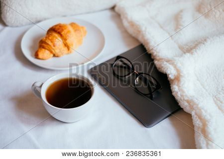 Big White Morning Cup Of Coffee And Crossant In Bed With Notebook, Glasses, Pillow And Cosy Blanket.