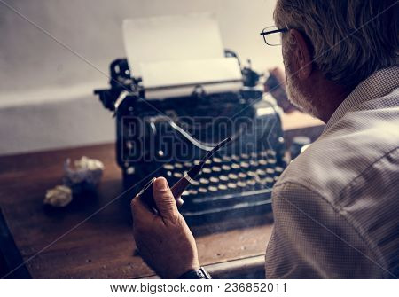 Elderly man sitting smoking pipe with retro typwriter on wooden table