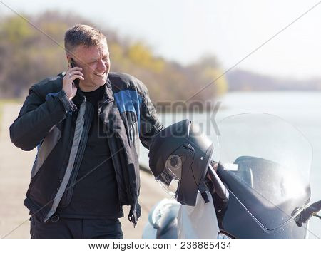 The Motorcyclists Is Standing Next To His Motor Scooter On The Road Near The Lake And Has A Conversa