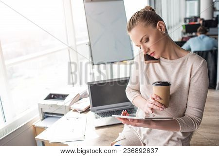 Beautiful Girl Is Sitting At The Adge Of The Table And Looking At The Tablet. Also She Is Talking On