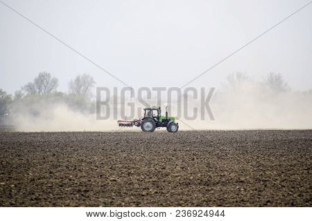 The Tractor Harrows The Soil On The Field And Creates A Cloud Of Dust Behind It.