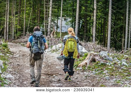 Rear View Of Two Young People Walking Down The Trail Path On Forest. Young Couple Hiking With Backpa