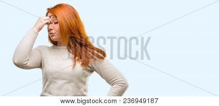 Beautiful young redhead woman with sleepy expression, being overworked and tired, rubbes nose because of weariness isolated over blue background