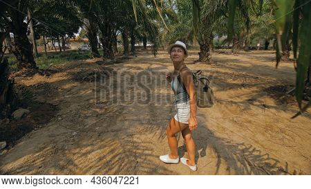 Woman Tourist With Plait Walks Looking Around At Growing Young Trees With Lush Leaves At Oil Palm Fa