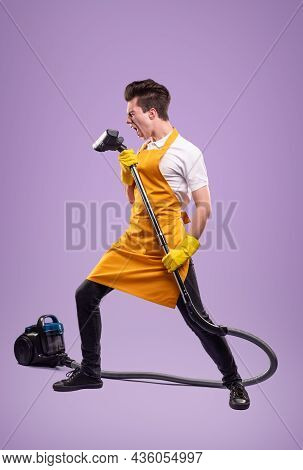 Young Househusband In Apron And Latex Gloves Singing Into Vacuum Cleaner While Standing On Purple Ba