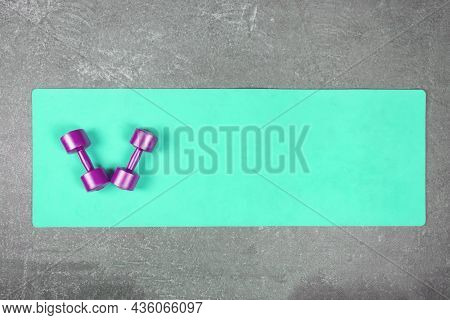 Flat Lay Of Turquoise Yoga Mat With Violet Dumbbells On Concrete Floor. Pair Of Dumbbells On Fitness