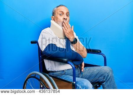 Handsome senior man with beard sitting on wheelchair with neck collar an arm sling covering mouth with hand, shocked and afraid for mistake. surprised expression 