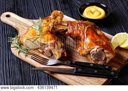 Crispy Baked Pork Shanks On A Wooden Board With Rosemary, Lemon Wedges And Mustard, Horizontal View 