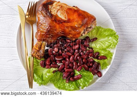 Crispy Roasted Pork Shanks On A White Plate With Fresh Lettuce Leaves And Baked Red Beans On A Woode