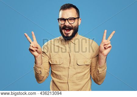 Optimistic Unshaven Man Screams And Shows V Sign Or Victory Gesture With Both Hands, Wears Eyeglasse
