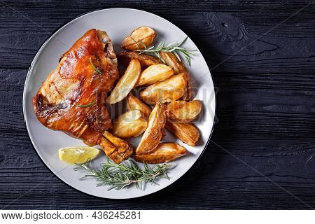 Crispy Roasted Pork Shanks On A Plate With Potato Wedges, Rosemary, Lemon On A Black Wooden Table, H
