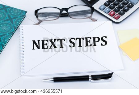 Next Steps Text On A White Notebook On An Office Desk Next To A Black Pen, Documens, Ring-up And Gla