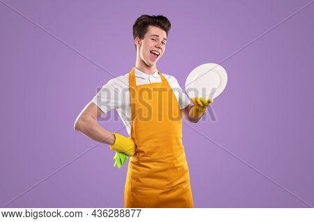 Satisfied Young Househusband In Apron With Hand On Hip And Plate Looking At Camera On Purple Backgro