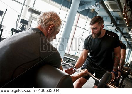 Back View Fit Senior Man With Gray Hair Sitting On Training Equipment, Holding Curved Barbell, Lifti