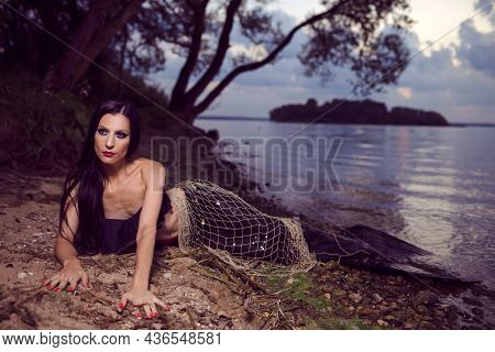 Mermaid Posing With Net Near Sea On Rocks While Wearing Artistic Makeup And Black Shiny Tail Covered