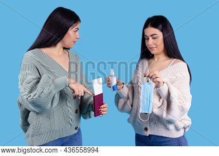 Two Twin Sisters On A Blue Background, Holding A Passport With An Airplane Ticket, Disinfectant And 