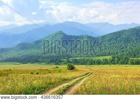 A Green Pasture Against The Background Of Mountains In The Morning Haze Under A Blue Cloudy Sky. Alt