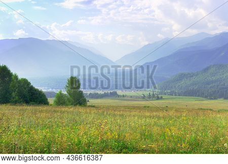 A Green Pasture Against The Background Of Mountains In The Morning Haze Under A Blue Cloudy Sky. Alt