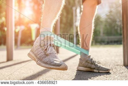 Closeup front view of legs feets of man guy training with elastic rubber band at the stadium outdoors. Athlete adult male person during active workout with additional sport equipmant in summertime