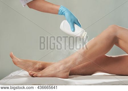 Cosmetologist is sprinkling and distributing talcum powder on a young girl's leg before the epilation procedure. The girl is lying on a couch in a beauty salon, she does the procedure shugaring