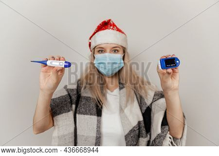Woman In Santa Hat, Wrap And Protective Medical Mask Holds Thermometer And Puls Oximetr