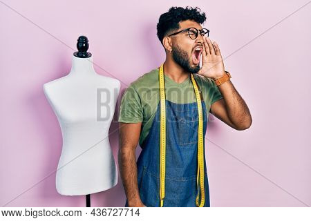 Arab man with beard dressmaker designer wearing atelier apron shouting and screaming loud to side with hand on mouth. communication concept. 