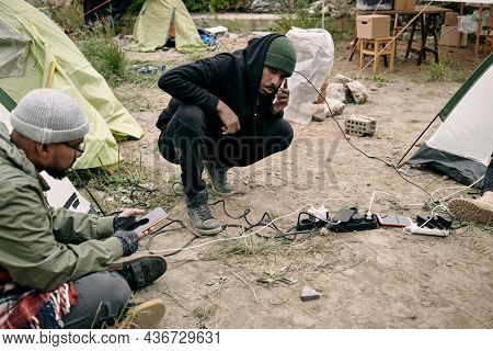 Two young male refugees using smartphones while sitting on ground in their camp