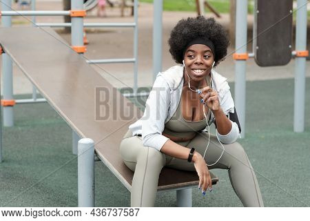 Young smiling sportswoman with earphones communicating through mobile technologies while sitting on sportsground