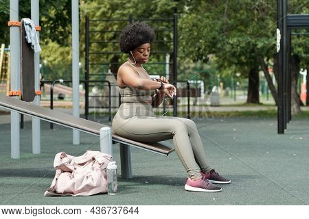 Plus size sportswoman in tracksuit looking at wristwatch while sitting on sports facilities