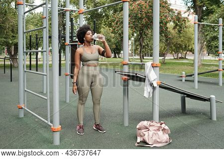 Young sportswoman standing on sportsground and having water during break after workout