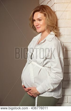 Beautiful Pregnant Woman With Blonde Curls And In White Blouse Holds Belly With Tenderness And Smile