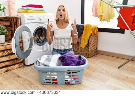 Young caucasian woman doing laundry with clothes in the basket amazed and surprised looking up and pointing with fingers and raised arms. 