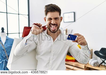 Young man with beard at retail shop holding shopping bags and credit card sticking tongue out happy with funny expression. 