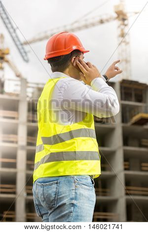 Young engineer in hardhat talking by phone and pointing at building site
